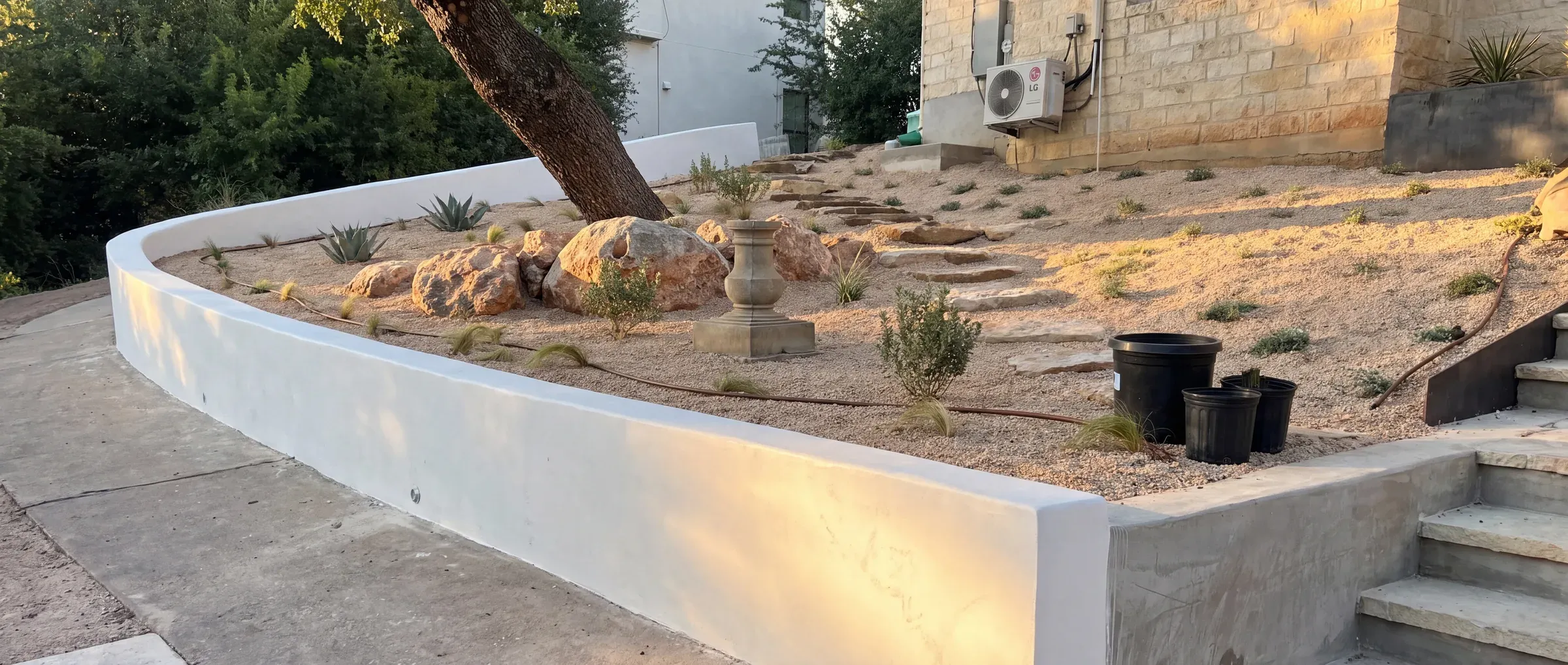 Planting day: a curved white retaining wall above a fresh bed of decomposed granite with boulders set, young plants going in, and nursery pots still standing on the gravel