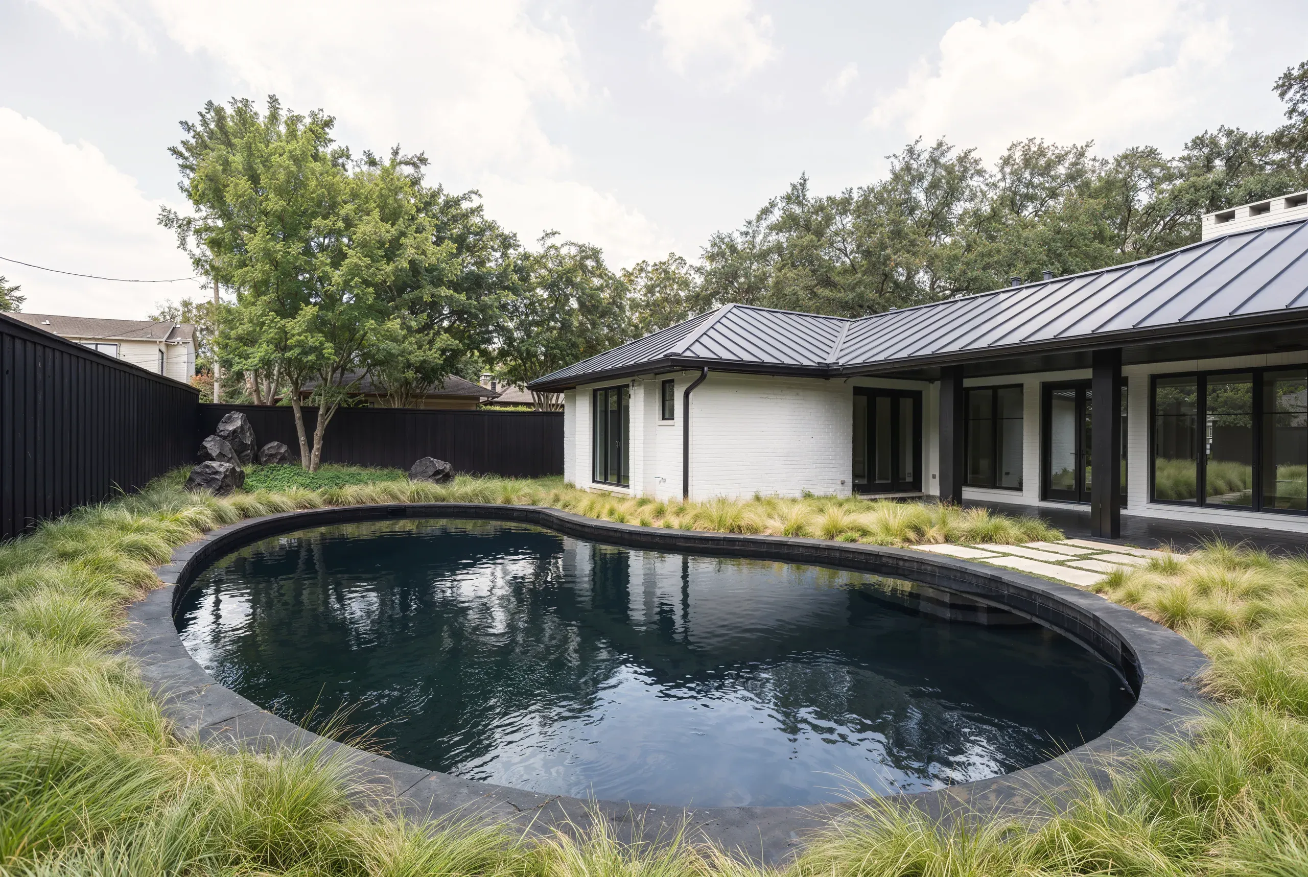 Austin backyard with a dark-bottom pool ringed in native grasses beside a white brick house under a black standing-seam metal roof