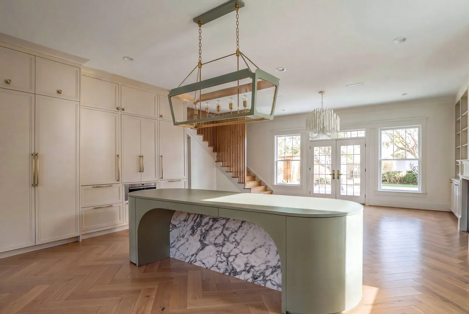 Cream French country kitchen with a curved sage green island on a marble base, herringbone wood floor and a green lantern pendant